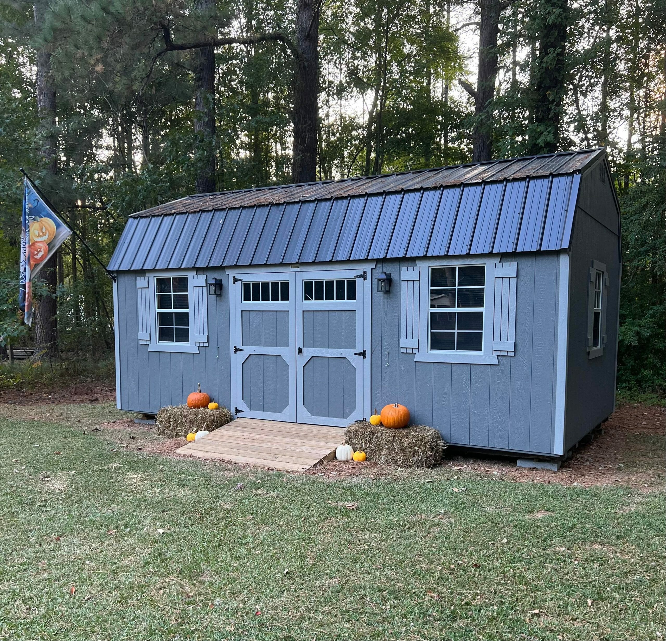 gray barn-style shed with a black metal roof is situated in a grassy yard surrounded by trees. The shed features double doors with decorative trim, two windows with white shutters, and outdoor light fixtures. It is decorated for fall with pumpkins and hay bales near the entrance. A festive Halloween-themed flag is displayed on a pole to the left of the shed.