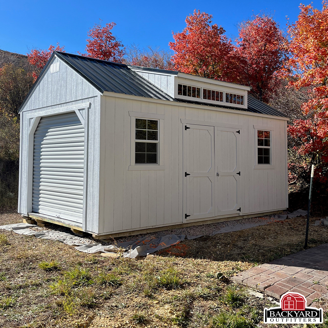 White utility barn with a dormer and roll-up garage door, surrounded by vibrant red autumn leaves and a clear blue sky.