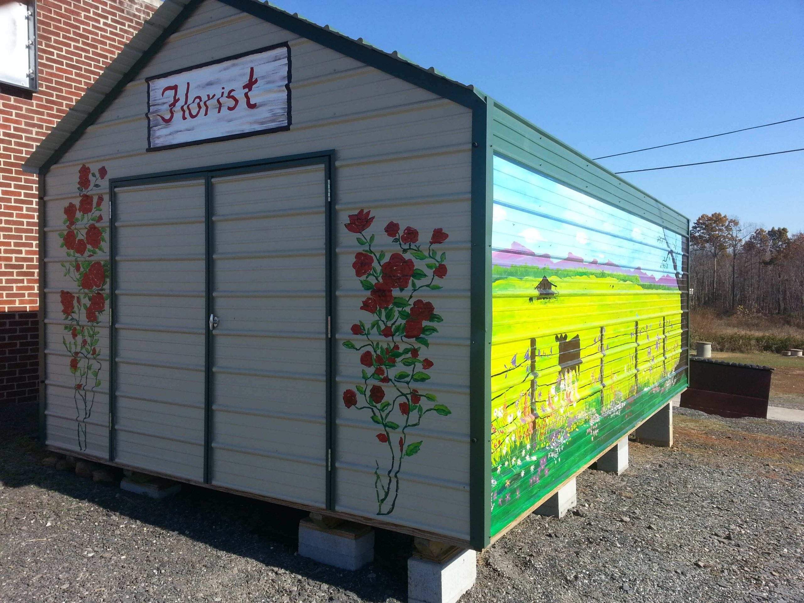 Light stone utility shed with hunter green trim, painted with a floral mural and a 'Florist' sign, set against a brick building and open landscape.