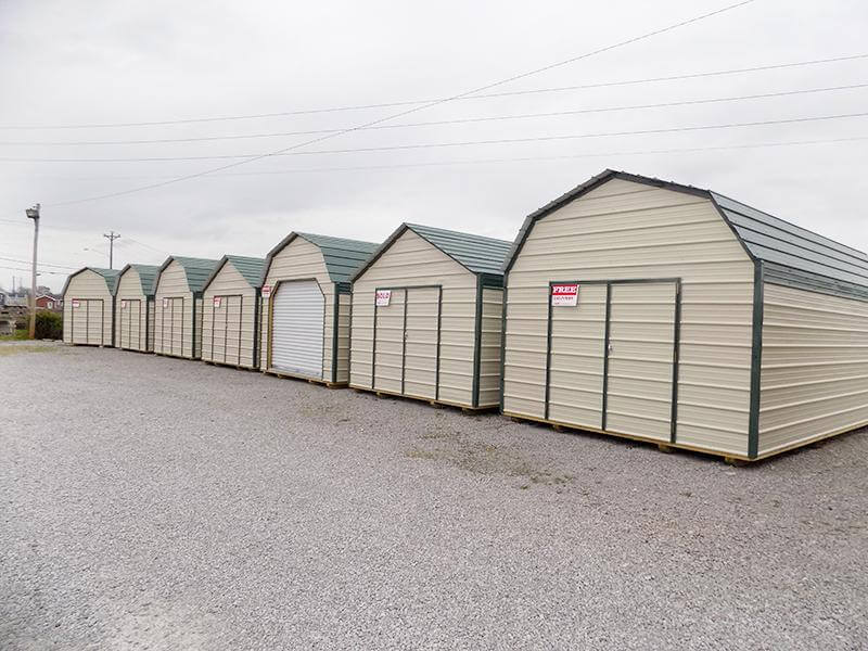 Row of metal lofted and utility sheds in light stone with hunter green trim, displayed on a gravel lot for sale.