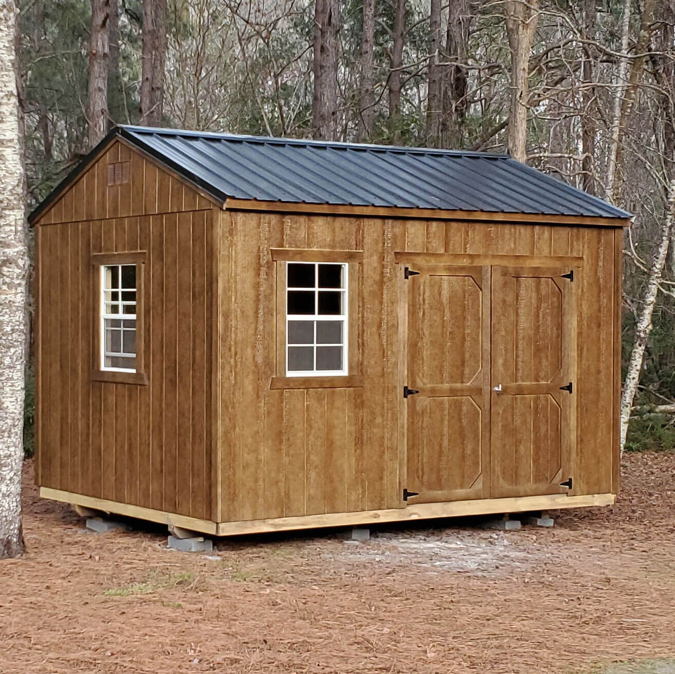 Rustic chestnut-colored utility shed with a metal roof and double barn doors, nestled among pine trees.