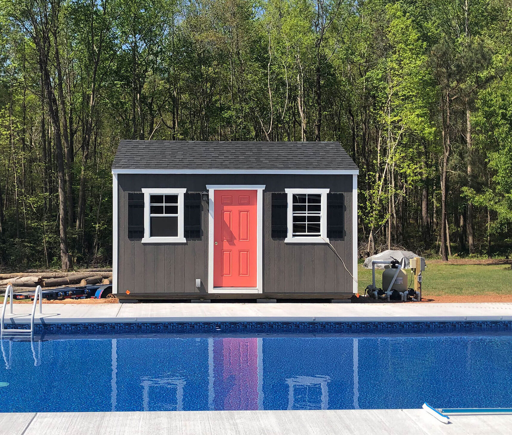 Gray utility shed with black shutters and a red door, located next to a pool and surrounded by lush greenery.