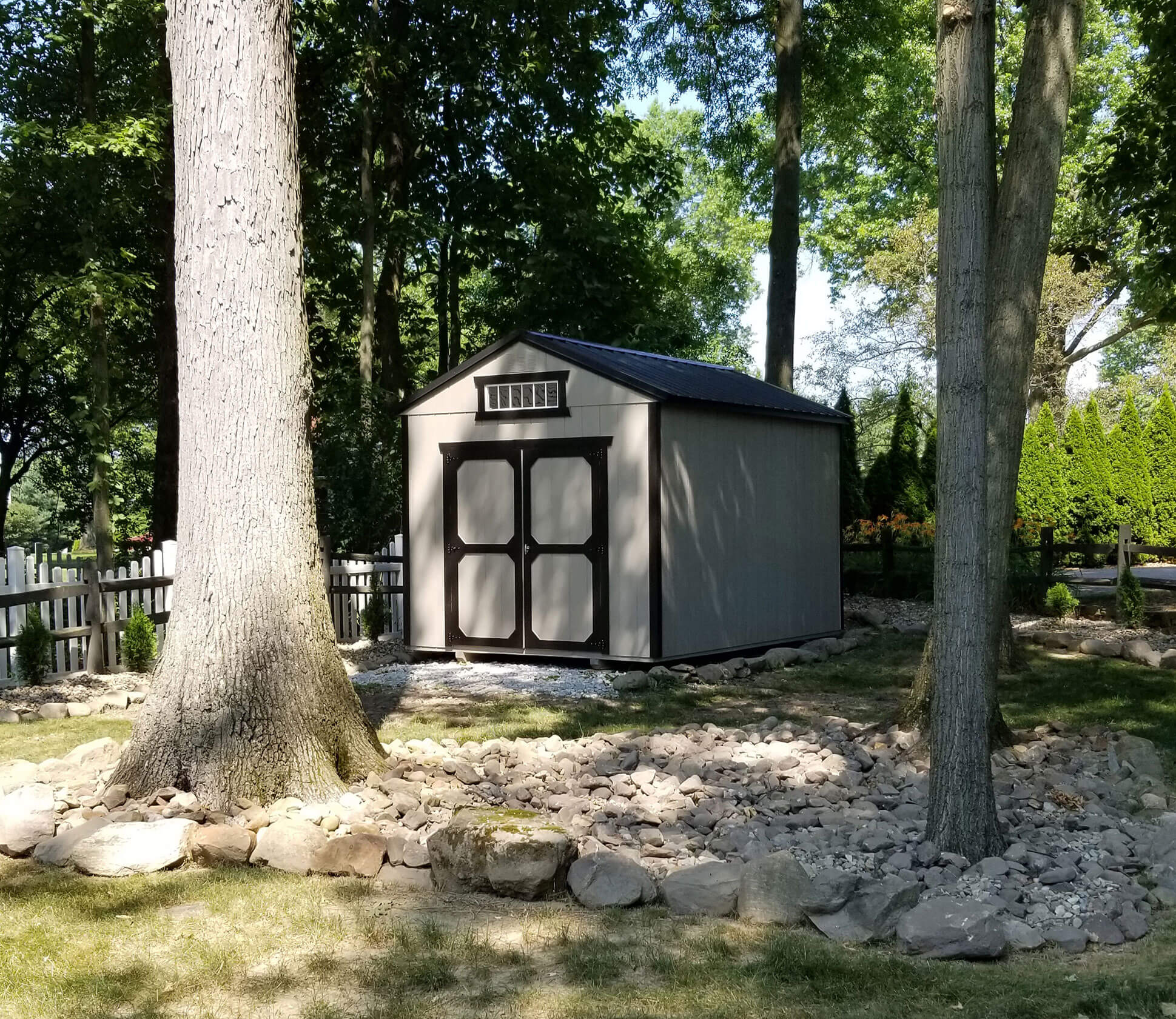 Gray utility shed with black trim and a transom window, situated in a landscaped backyard with large trees and a rock garden.